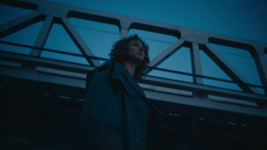 A woman standing below a bridge nervously turns around and looks behind her