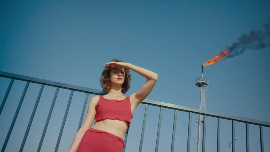 A woman wearing a red halter top and red pants is standing in front of a fence with a flaming torch in the distance behind her