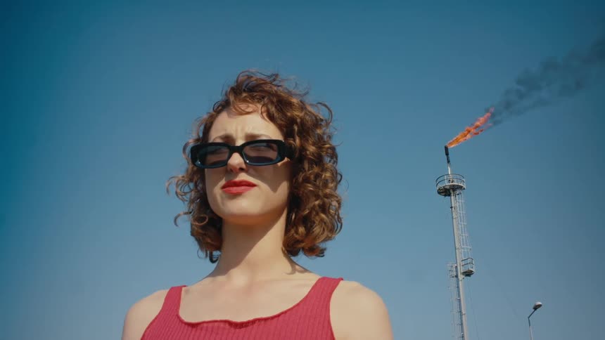 A woman standing in front of a large tower with a flame coming out of the top adjusts her sunglasses
