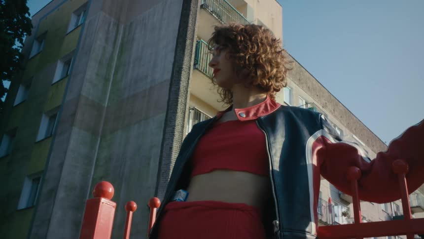 A woman wearing red clothes and a jacket is standing on a street leaning against a red fence and looking around and there is an apartment building behind her