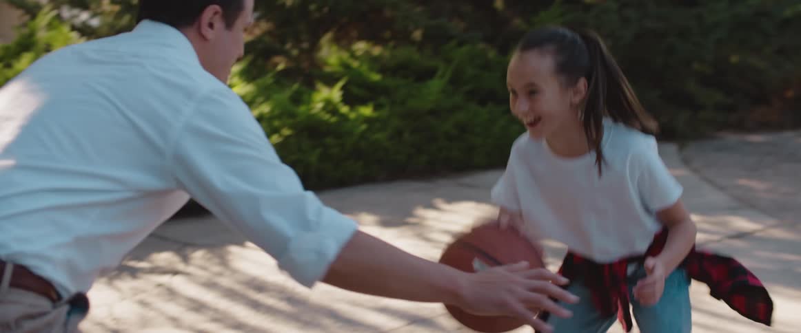 A girl playing basketball outdoors against her dad dripples for a moment before shooting the ball over his head and cheering