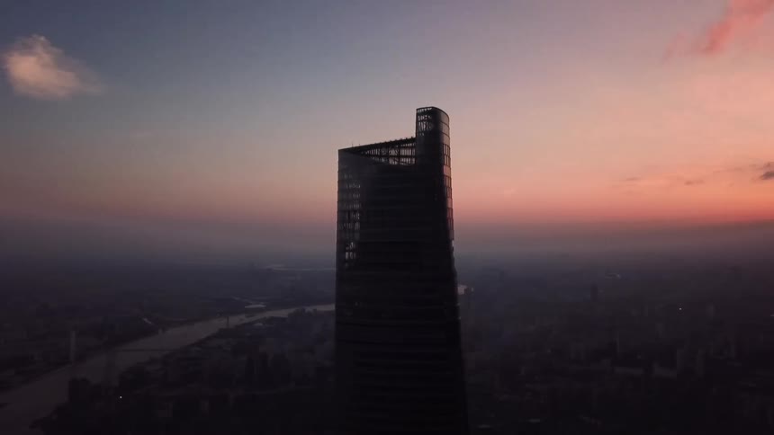 A skyscraper overlooks a river and a city in a hazy colorful sky