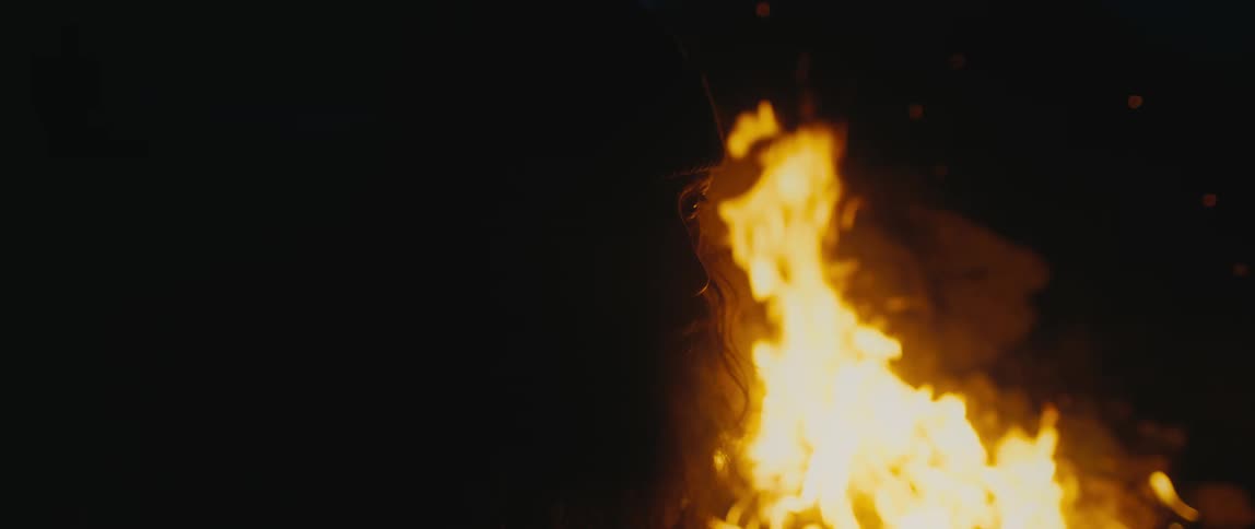 A man with long hair and a beard is in the darkness as a bonfire is burning brightly behind him