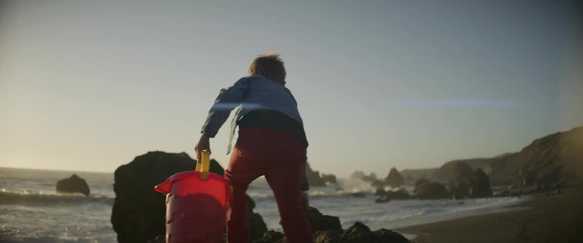 Young boy carries a large red pail and a stick as he climbs over the rocks near the ocean