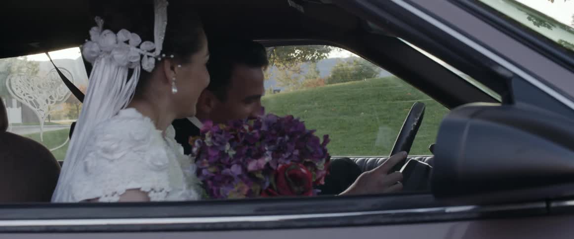 Groom in tuxedo gets in car next to bride with wedding dress holding a bunch of flowers
