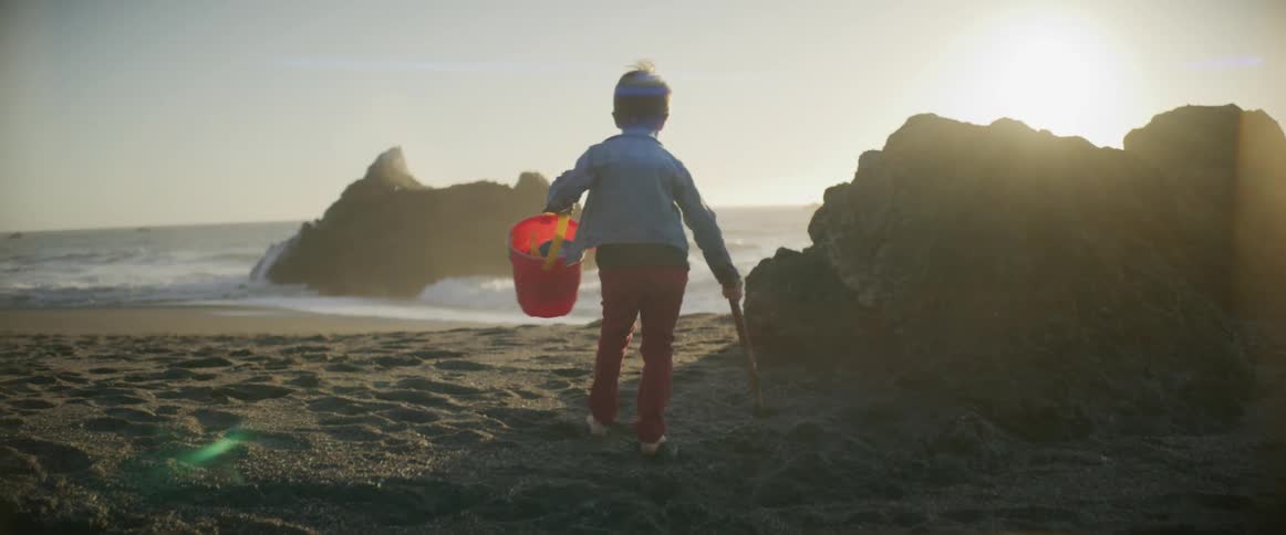 Young boy wearing red pants carries a red pail and a stick as he walks towards a large rock on the beach near the ocean