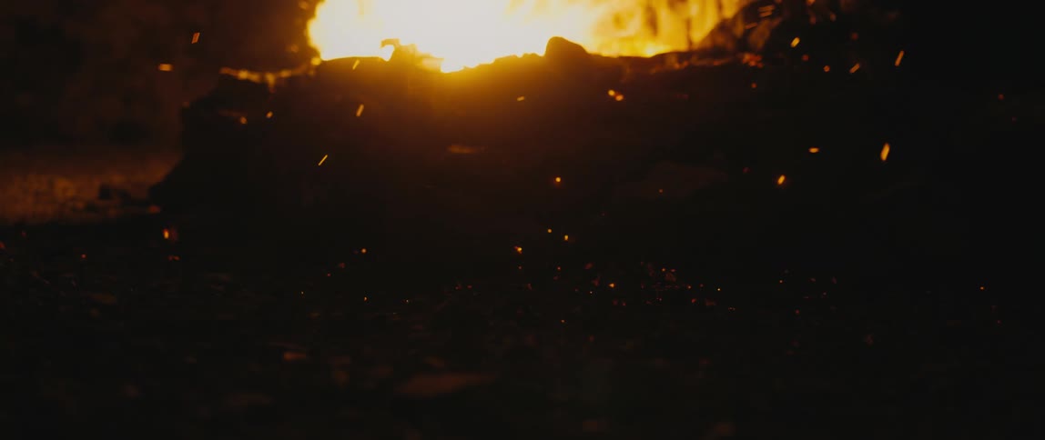 Embers emitting from a campfire fall onto the surrounding soil at night