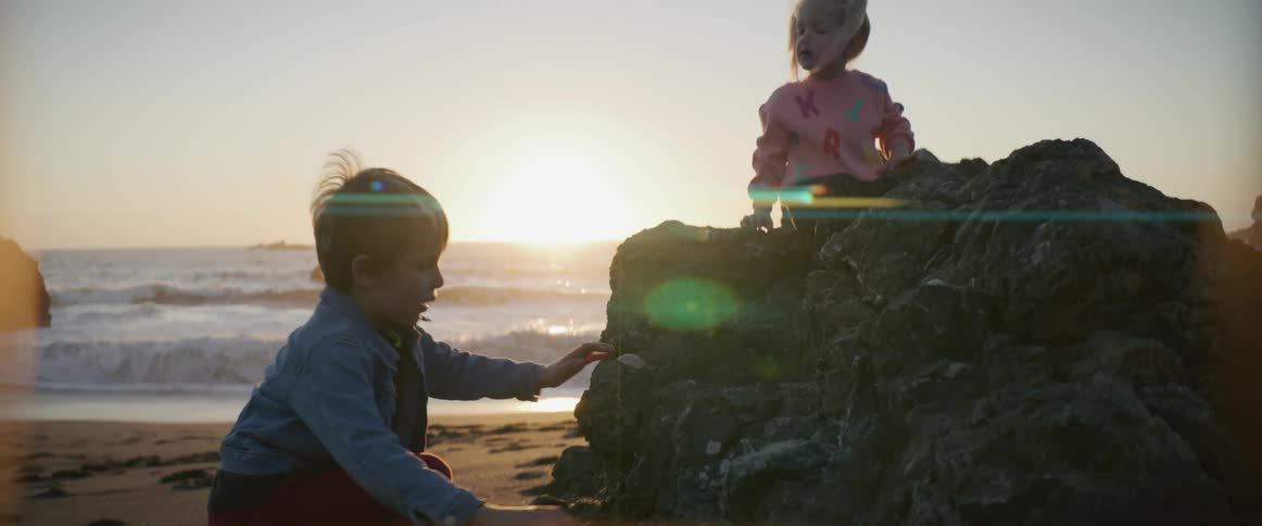 A boy climbs a rock formation on a beach to reach a girl already sitting at the top