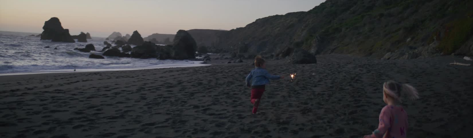 Little boy carries a sparkler as he runs on the beach and his sister follows him
