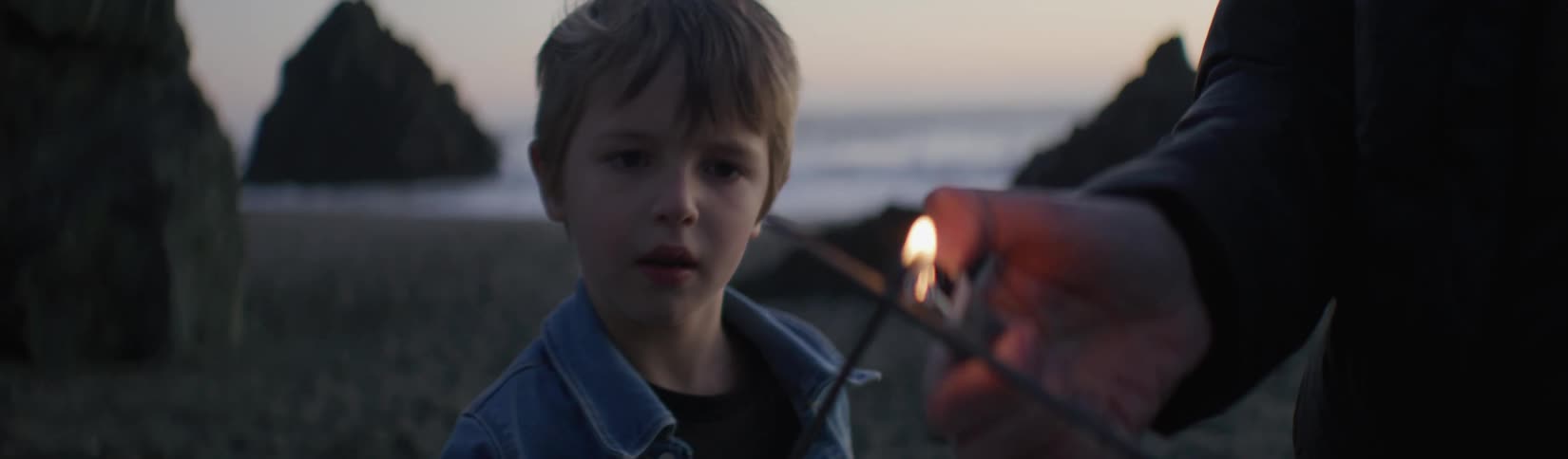 A dad uses a lighter to light the sparklers he and his son are holding at the beach