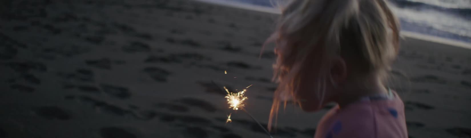 A young girl runs on a beach at dusk with a sparkler in her hand