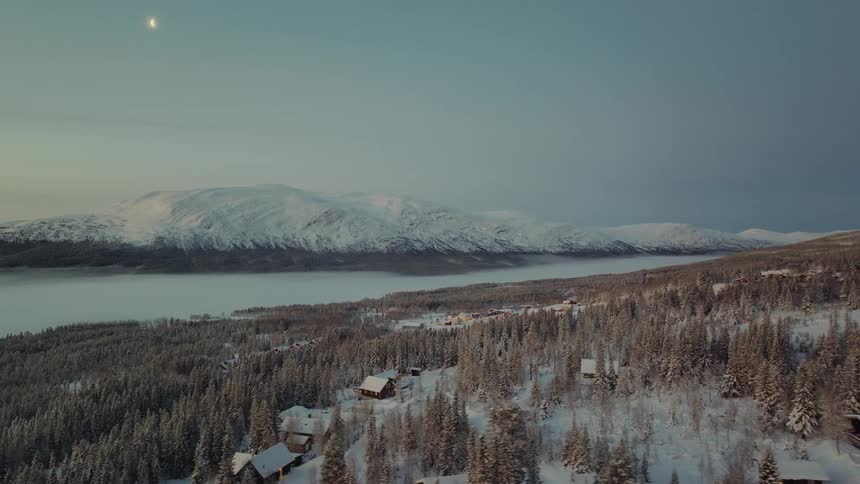Sliver of the moon sits in the sky over snow covered mountain sitting across water from town and forest