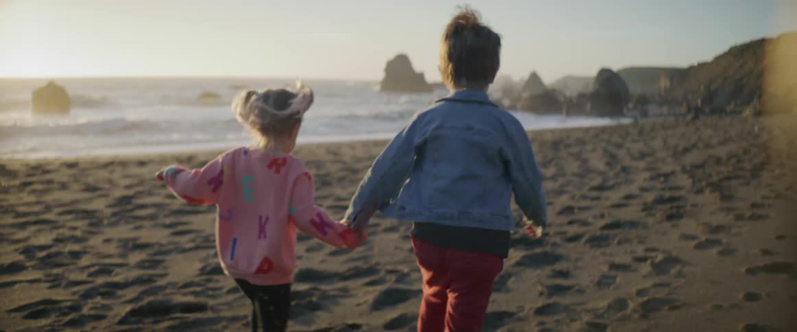 A toddler holds her brothers hand as they run on the beach
