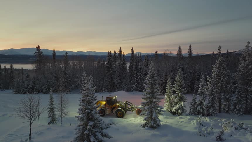 A snow plow plows snow in a forest area in front of a lake