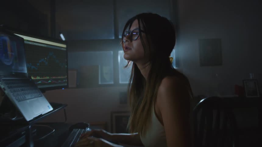 A longhaired woman wearing spectacles sits in a dark room in front of a window focusing on several large computer screens while typing on a keyboard
