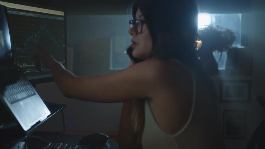 Woman working in office at night talks on phone while examining data on screen