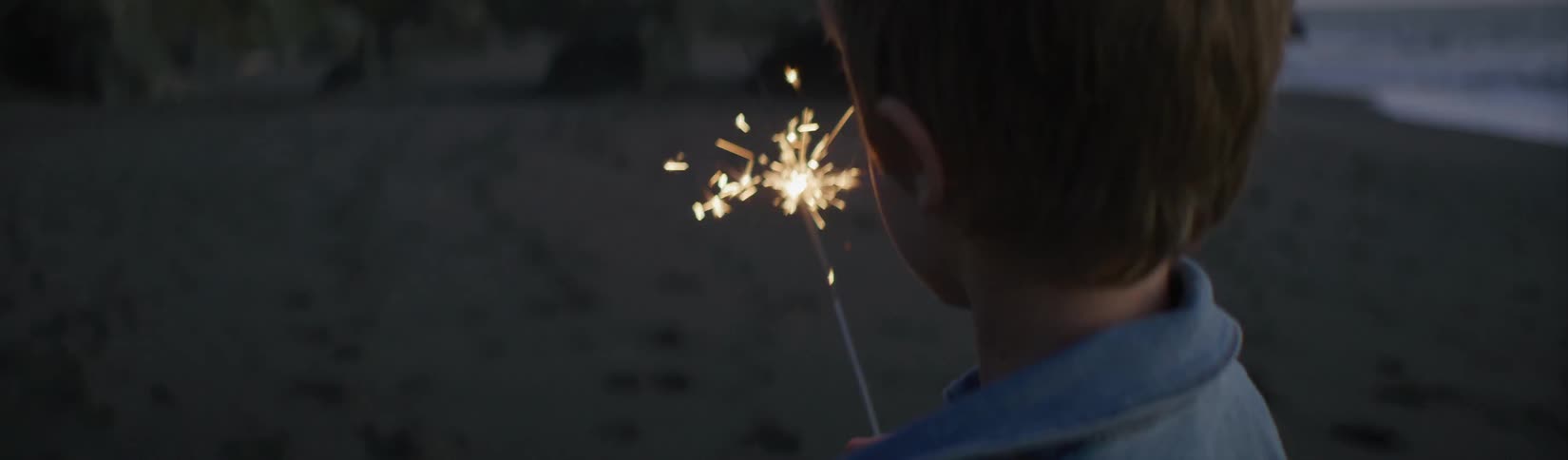 A young boy standing on a beach watches a sparkler burn as his dad and sister walk towards him