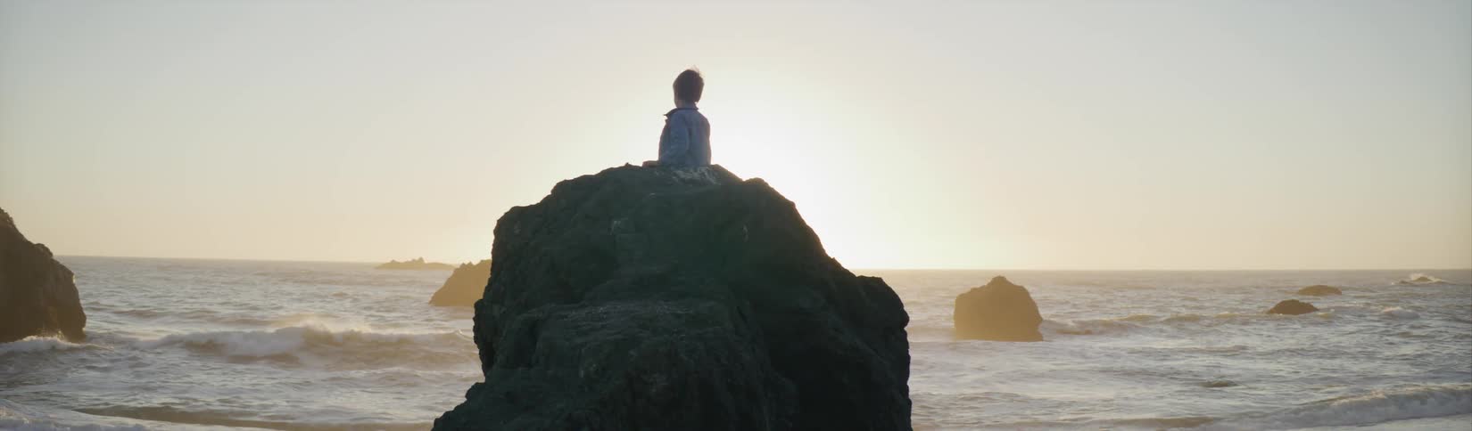 A young boy sits on a large rock on the shore and looks at the waves rolling in