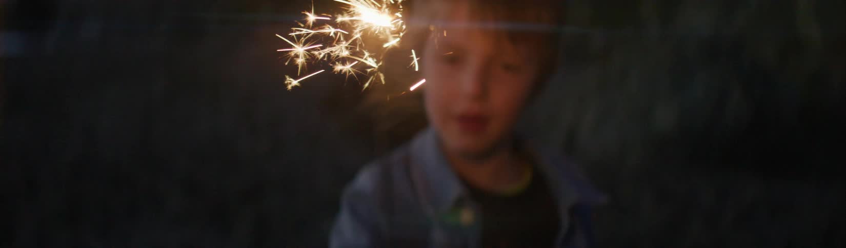 A young boy is on a beach holding a sparkler and smiles as he waves it around