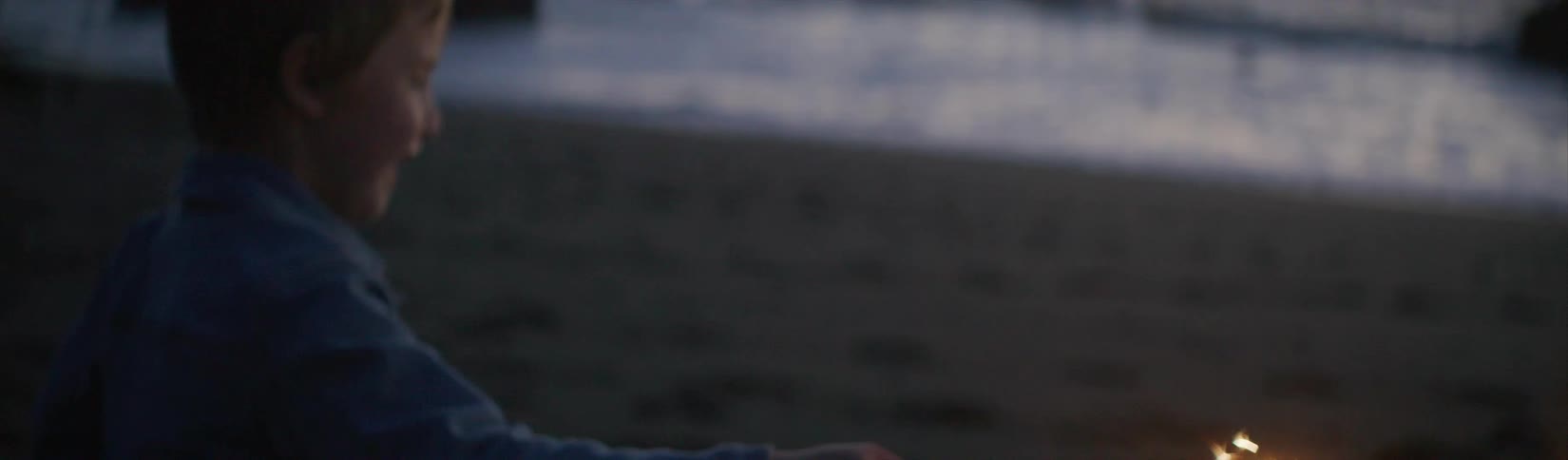 A young boy walking on a beach laughs as he holds a sparkler and watches it burn