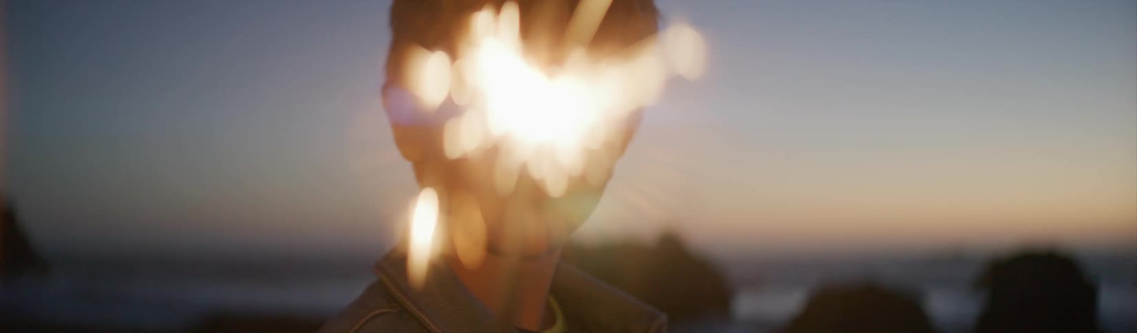 A young boy stands on the beach in the evening and watches a sparkler he is holding burn