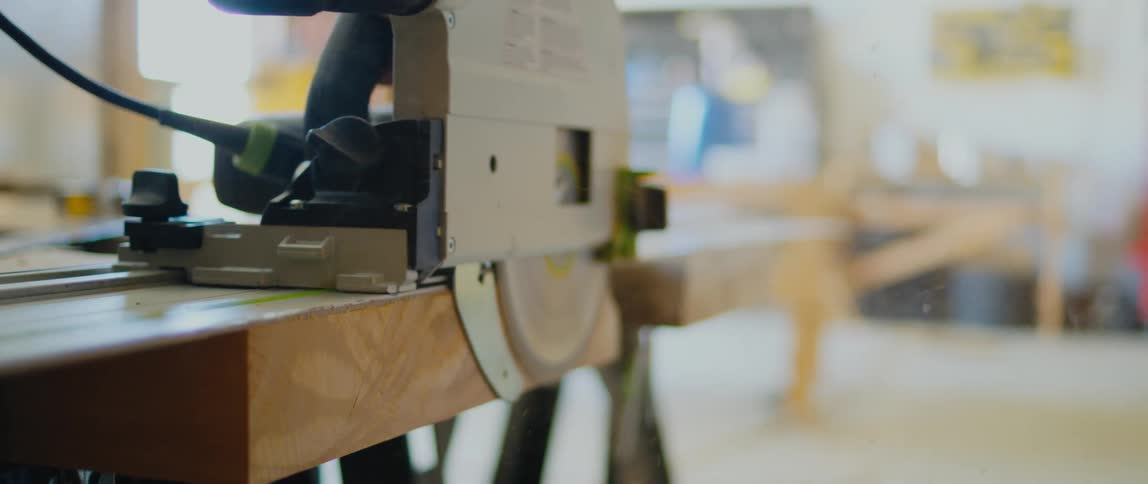 Man trims the edge of a flat piece of wood over a workbench using a circular saw inside a workshop