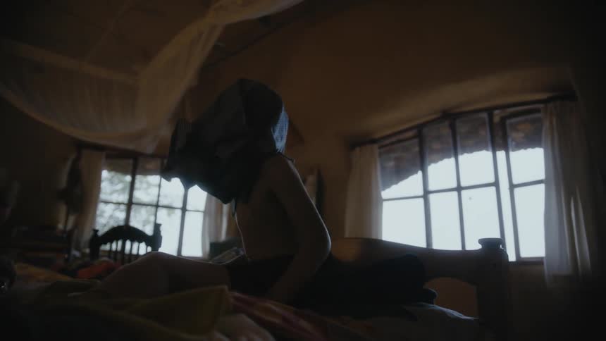 A woman sits on the bed with her son and laughs as he puts on a shirt and keeps it over his face and looks around