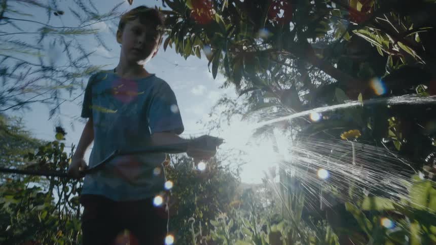 A boy waters flowers and plants with a garden hose as his mother walks behind him