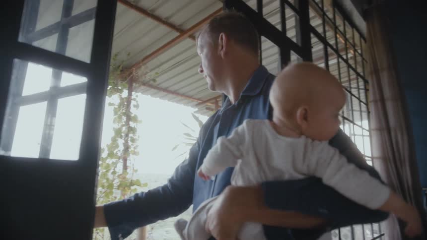 Father with blue shirt carries baby and goes out of house in the countryside