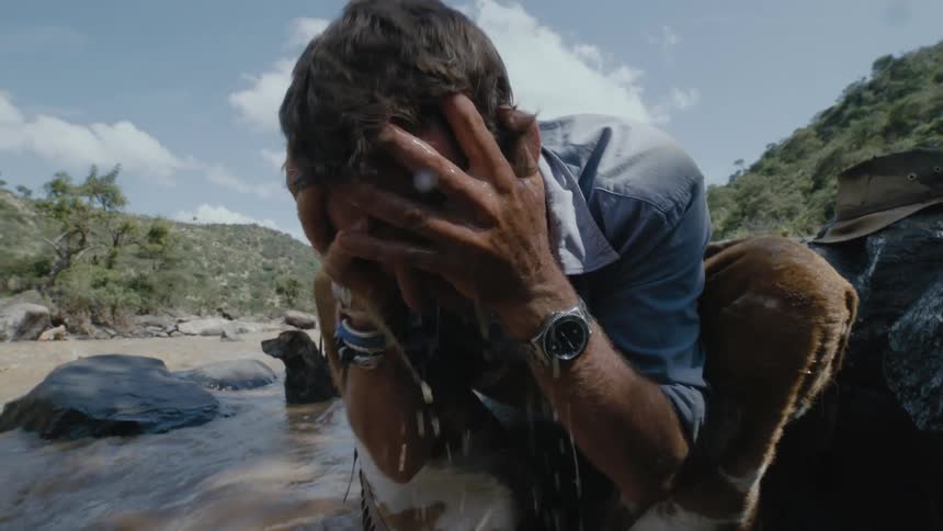 A dog sits in a river while his own puts river water in his hands and drinks it and then splashes it all over his face and hair