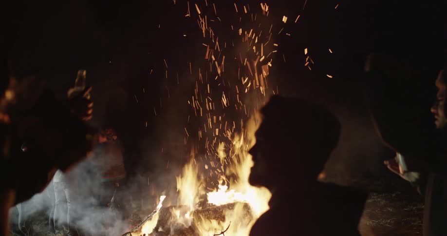 A group of people dance, drink, and sing around a bonfire