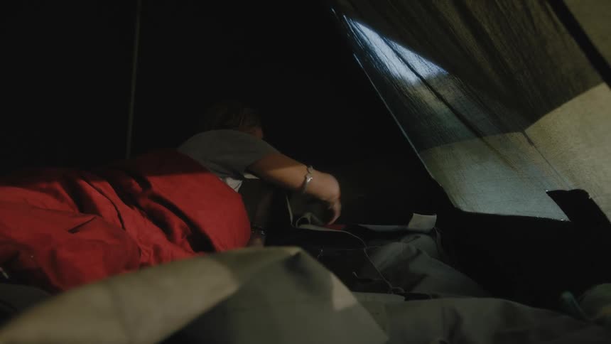 Young woman laying inside a tent ties the laces on the sleeping bag of her companion at morning