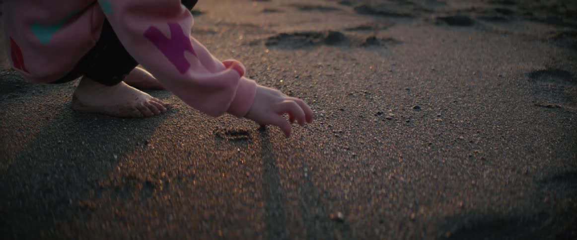 Blonde girl with pink sweater and black pants crouches down and draws on sand on the beach at sunset