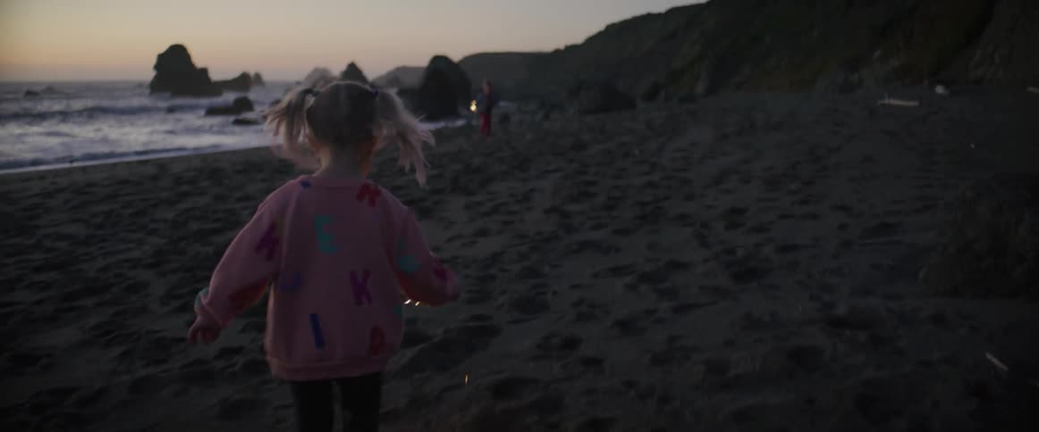 Little girl holding a sparkler firework stick running in the beach next to the ocean towards her brother