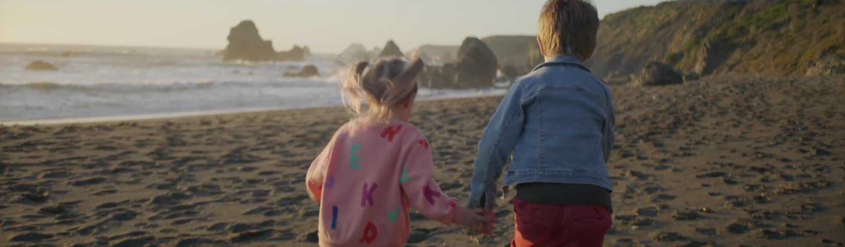 A brother and sister hold hands while running on a sandy beach under the sun
