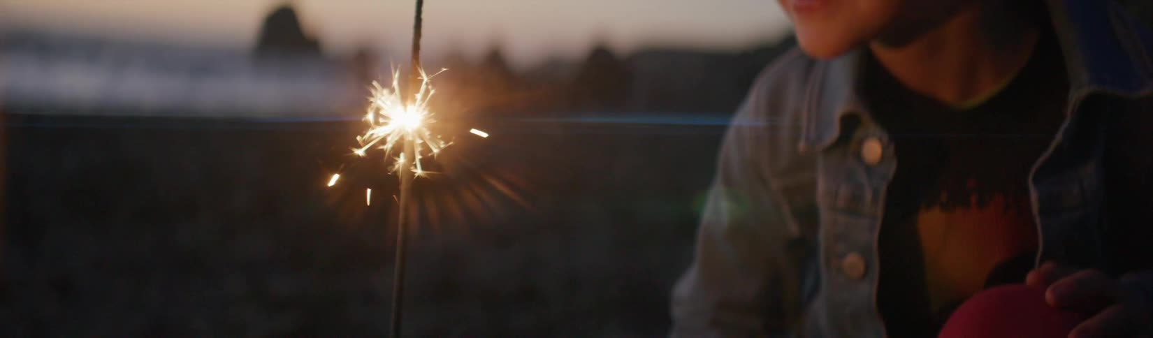 A young boy is on a beach holding a sparkler and smiles as he watches the sparks