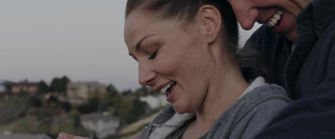A woman smiles and admires her engagement as her fiance hugs her from behind