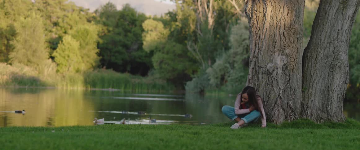 Young girl reading a book under a tree near a lake at morning stands up and walks away