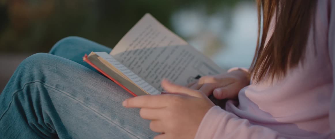 Girl with brown hair pink sweater and jeans reads book by the lake