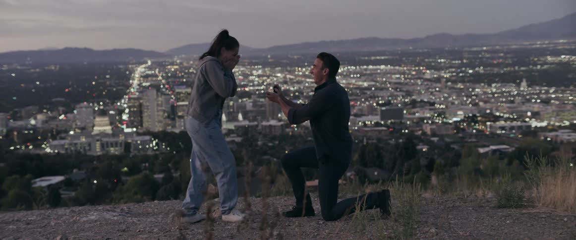 Man kneels on the ground to propose to his girlfriend over a hill in front of a small town at evening