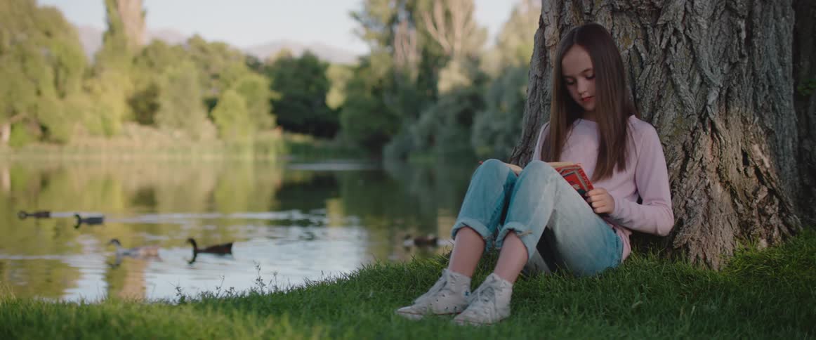 Teenage girl sitting against a tree trunk next to a lake reading a book at a sunny day