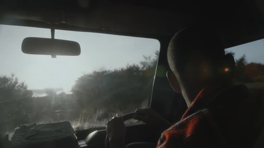 Black man driving a car through a forest area with sunlight entering the car through the front window