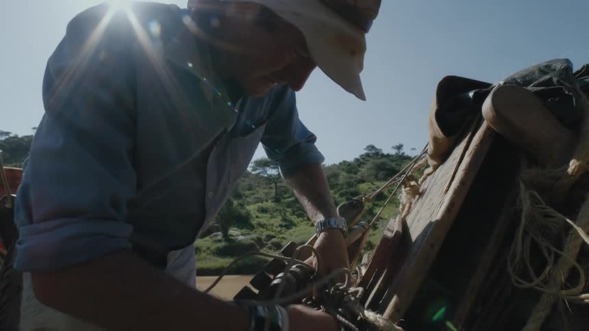 Man wearing a sun hat uses rope to tie objects to the back of a car outdoors