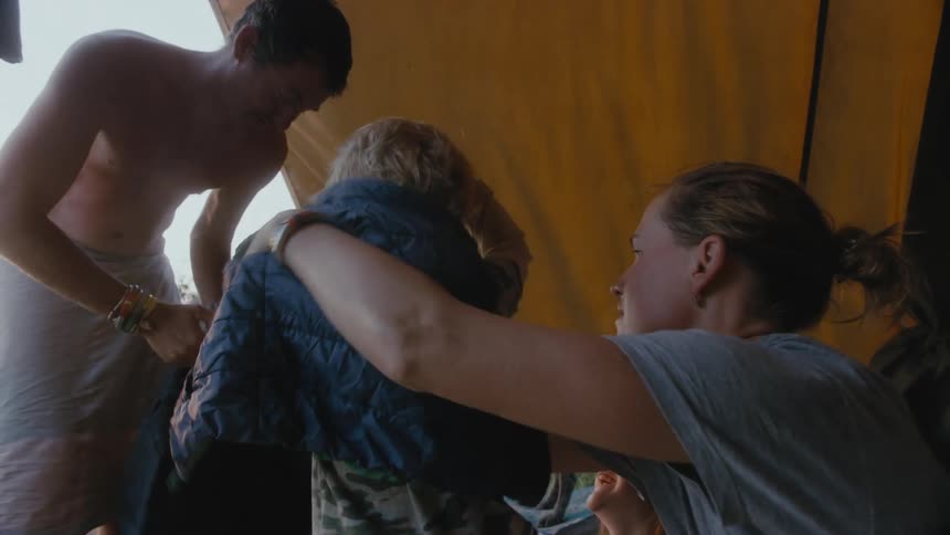 Mother putting jacket on her son under a yellow camping tent while the father is putting on a short
