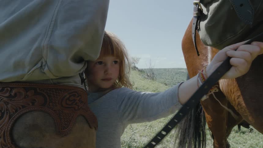 Young blonde girl holds a leash hanging from a horse saddle while an adult helps her put a helmet on