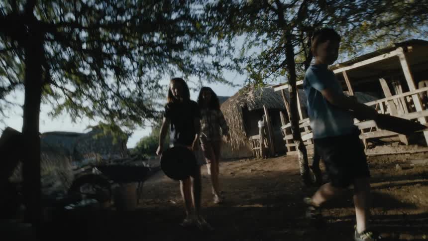 Brother and sister each carry a bowl while their mother follows them as they walk past a hut