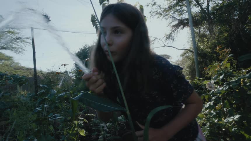 Girl with wavy hair and dark blouse waters the garden and drinks water from hose