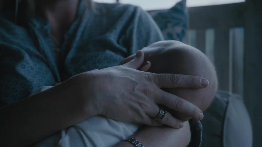 Mother caressing the face of an asleep baby on her hands while sitting on a couch at home