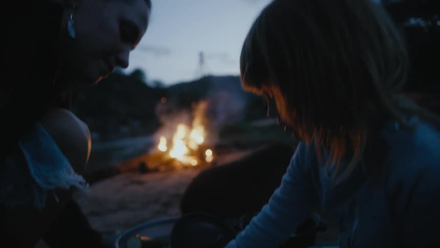 A woman cuts food and her young daughter puts it in a pan as they sit outside by a fire