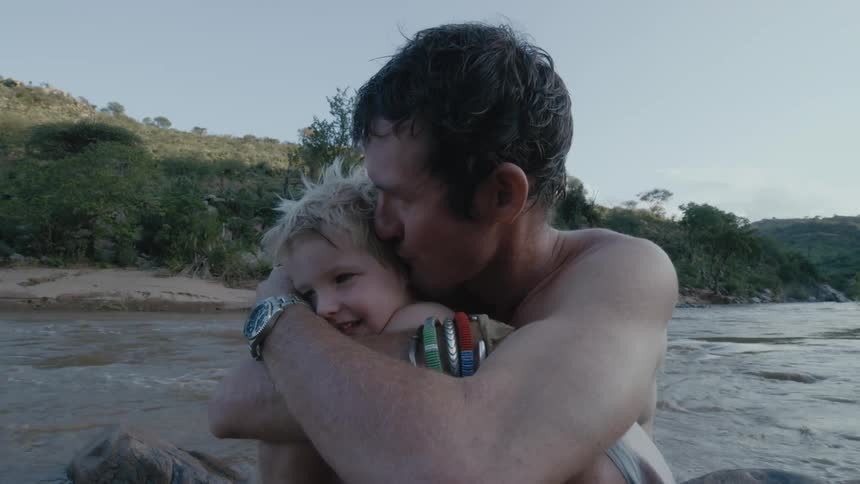Shirtless father hughes his son from behind while sitting next to the river